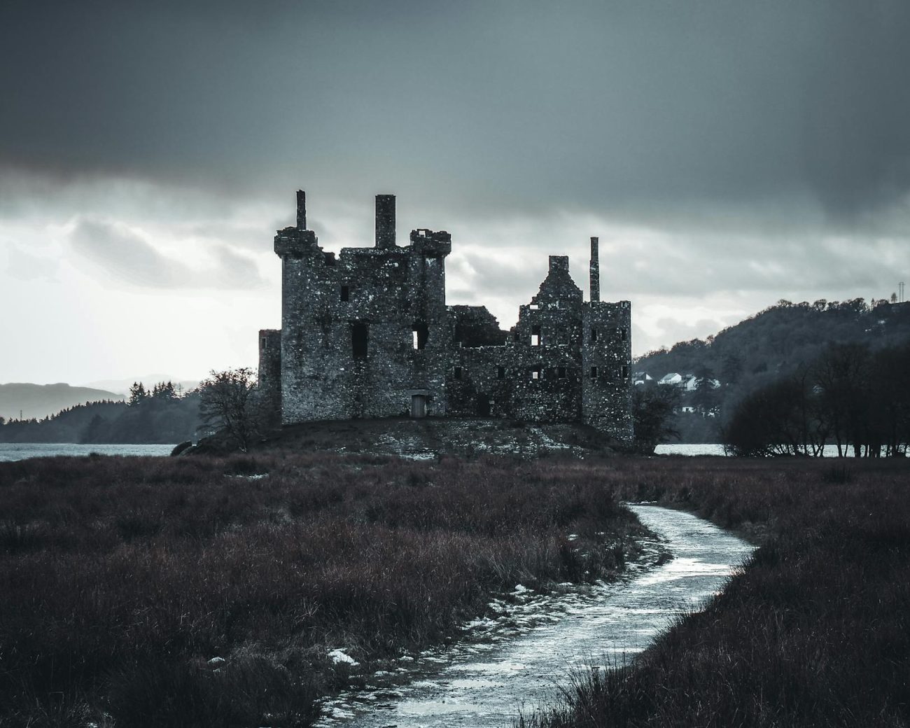 A historic Scottish castle sits by a lake under a dark, stormy sky, creating a moody atmosphere.