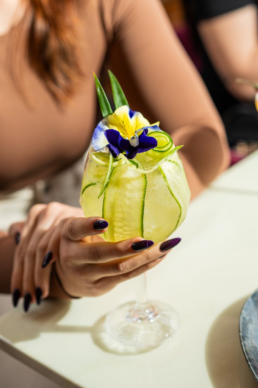 Hand holding a floral cocktail garnished with cucumber and flowers. Perfect for a summer gathering.