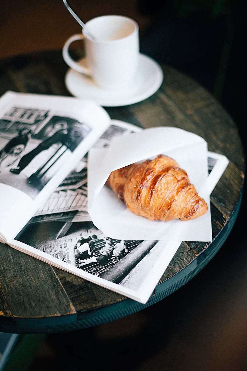 A cozy setup featuring a warm croissant and coffee cup on a wooden table with an open magazine.