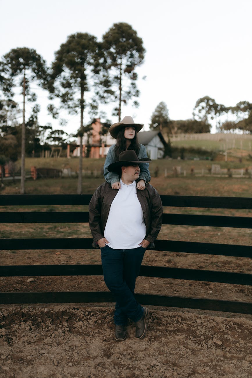 Couple wearing hats, leaning on fence at a rustic farm with trees and house in background.