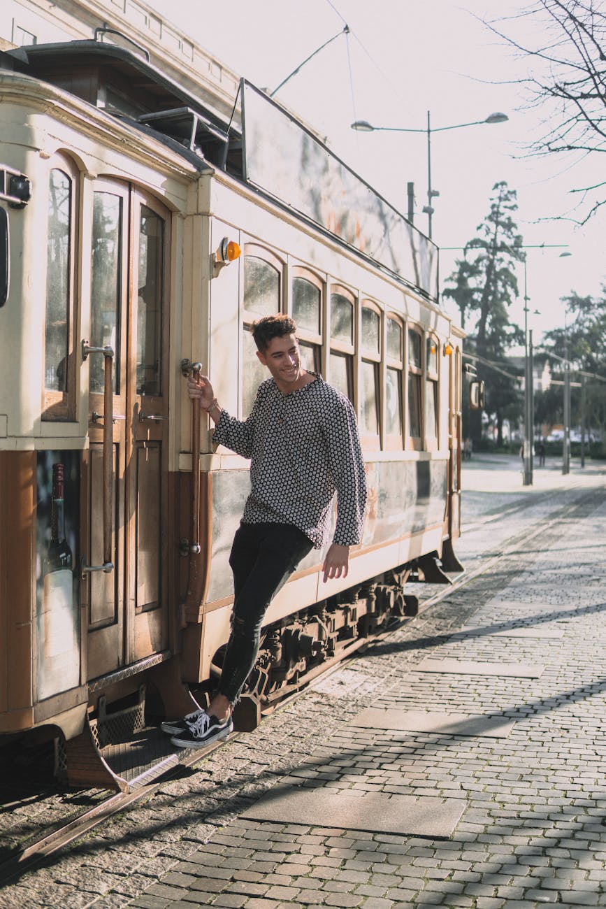 A young man playfully rides an old tram in a sunlit city street.