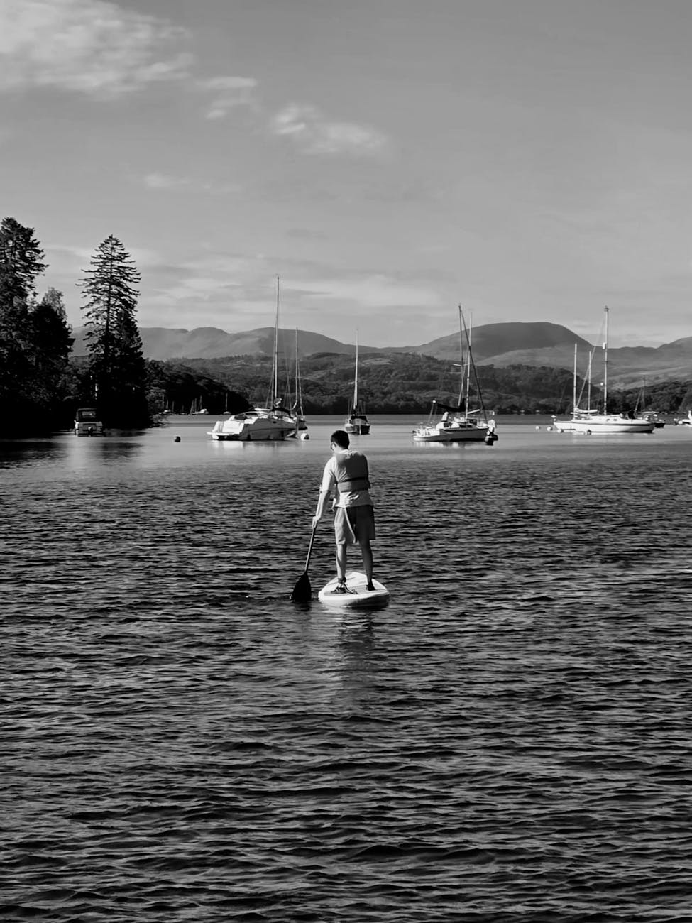 Person paddle-boarding on a tranquil lake surrounded by sailboats in a mountain landscape.