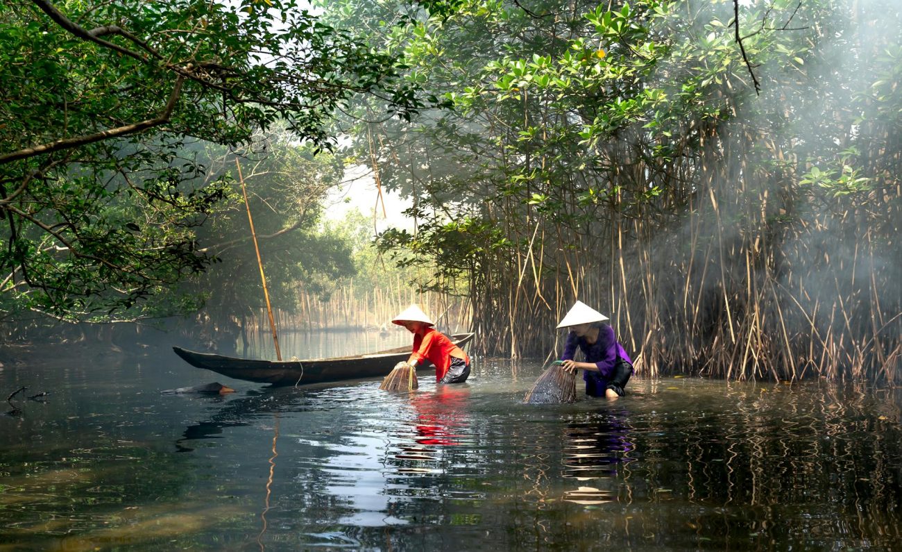 Two women using traditional bamboo baskets to fish in a serene river setting.