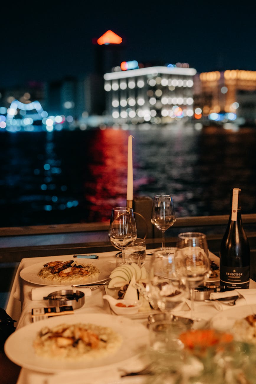 Enjoy a romantic dinner by the river at night, featuring elegant plates and a stunning cityscape view.