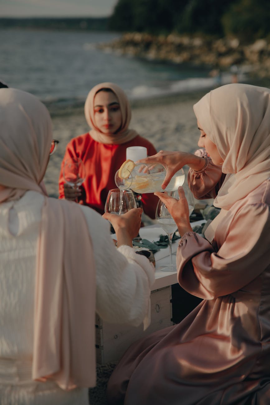 A group of women sharing drinks and enjoying a sunset picnic by the shore, dressed in hijabs.