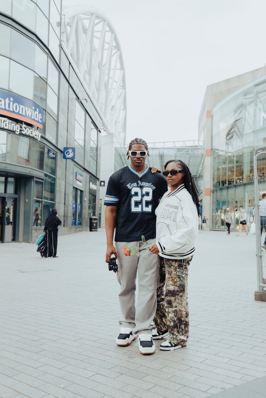 A fashionable couple with trendy outfits and sunglasses is posing in a modern urban downtown setting.