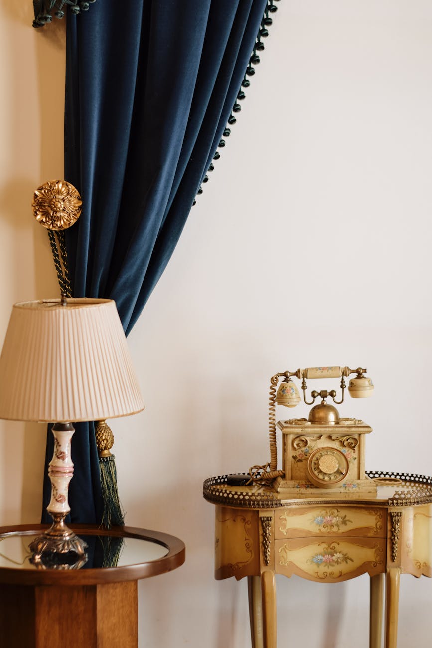 Elegant vintage room setup featuring an ornate antique telephone and classic table lamp beside a blue velvet curtain.